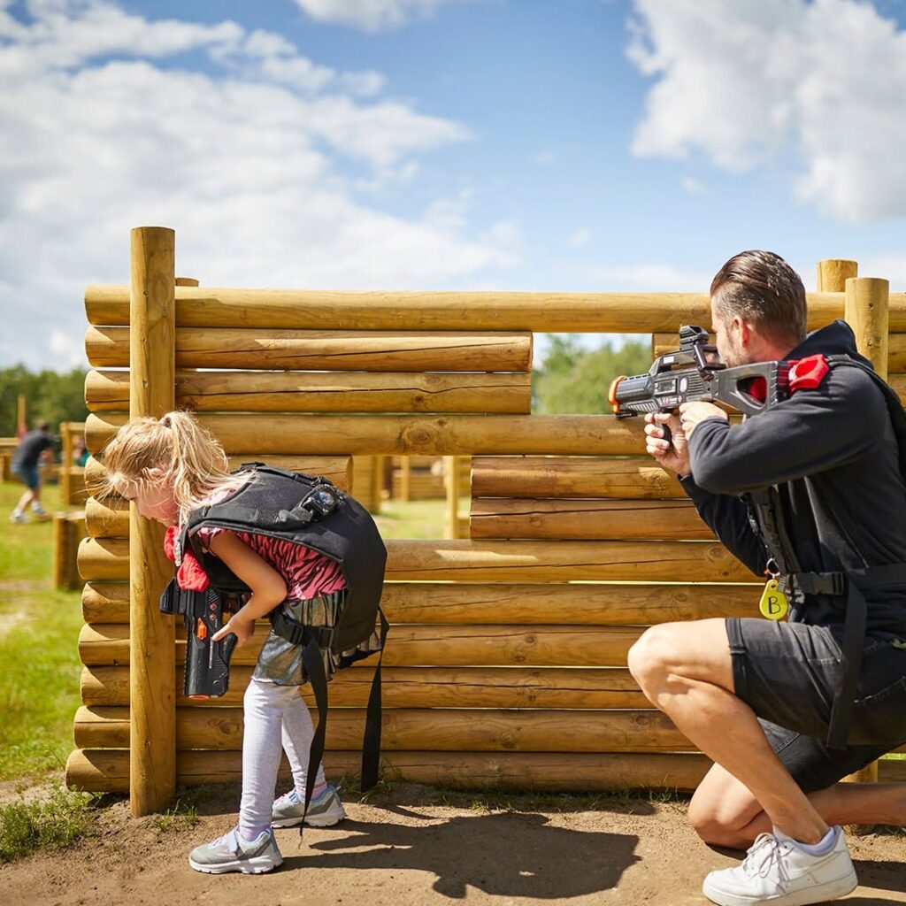 Vader en dochter spelen samen een outdoor lasergame op een houten speelveld tijdens een actieve gezinsactiviteit.